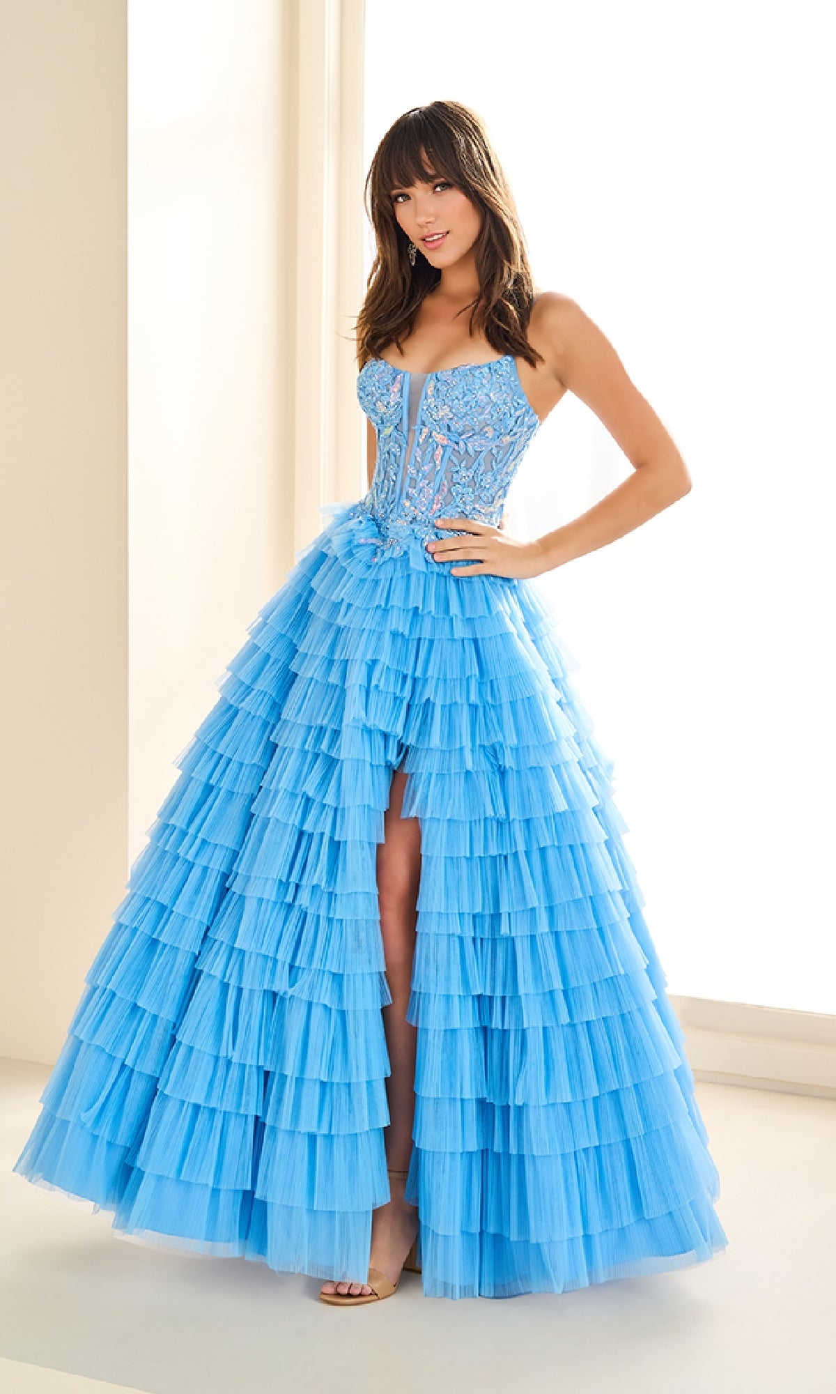 A woman stands indoors wearing the Ellie Wilde EW36086, a long strapless prom dress with a corset bodice and layered blue tulle skirt.